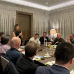 An engaged group of Fractional Chief Financial Officers (CFOs) in a collaborative meeting, with a female speaker leading the discussion at the head of an elongated conference table. The setting is a stylishly decorated room with star-patterned wallpaper, suggesting a formal yet dynamic exchange of ideas among experienced finance professionals.
