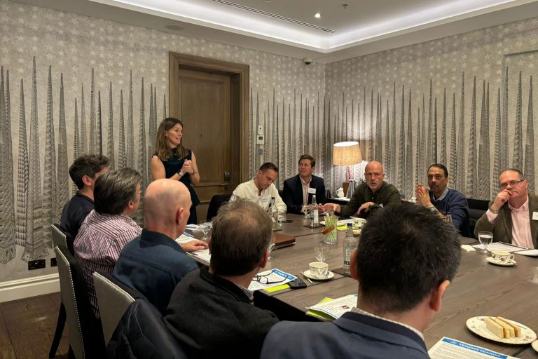 An engaged group of Fractional Chief Financial Officers (CFOs) in a collaborative meeting, with a female speaker leading the discussion at the head of an elongated conference table. The setting is a stylishly decorated room with star-patterned wallpaper, suggesting a formal yet dynamic exchange of ideas among experienced finance professionals.